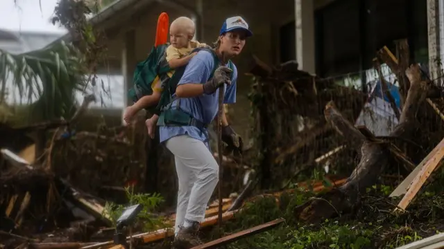 A woman walks through wooden debris outside a damaged home, a small toddler in a yellow shirt held in a backpack on her back