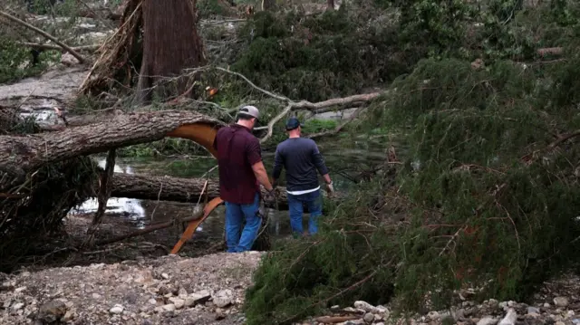 A man (L) in short-sleeved, burgundy shirt and jeans walks with another man (R) in long-sleeved, blue shirt and jeans towards what appears to be a river. They are walking among fallen trees as they search for bodies and survivors following flash flooding
