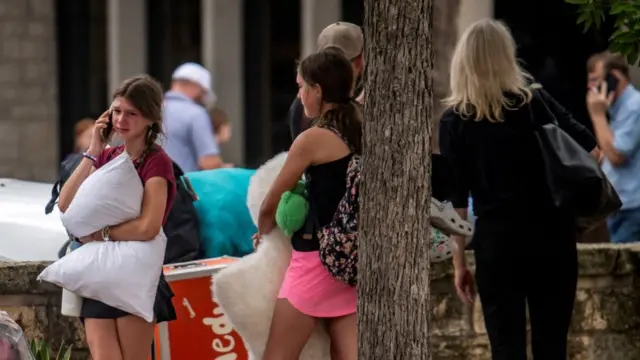 A young girl in tears holds a pillow while on the phone, as others wait nearby