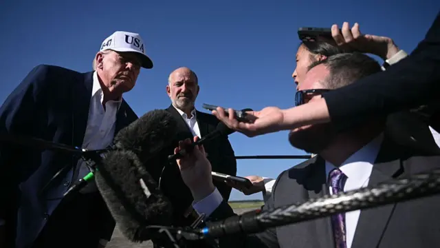 Donald Trump in white USA cap answers questions from the press while on the tarmac, Secretary of Commerce Howard Lutnick standing to his left in a suit