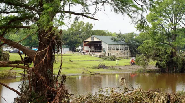 Wide shot of main wooden building at Camp Mystic with missing front. Shot is taken from across the river, showing water flow by and people on the grass