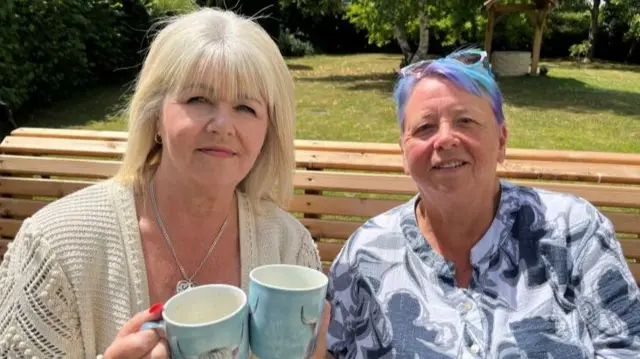 Nichola Arch (L) and Wendy Buffrey (R) holding cups of tea on a wooden bench