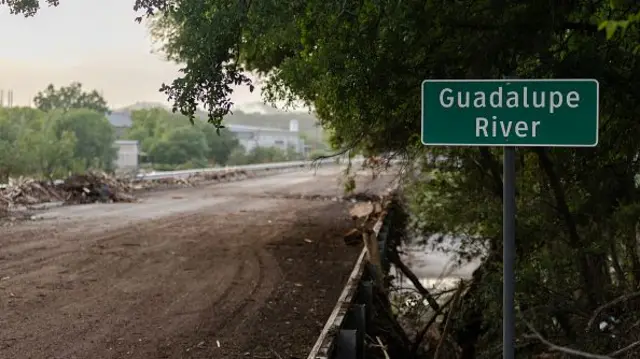 A bridge that spans the Guadalupe River with a green sign showing the name of the river. Brown debris and uprooted trees fill the sidewalks