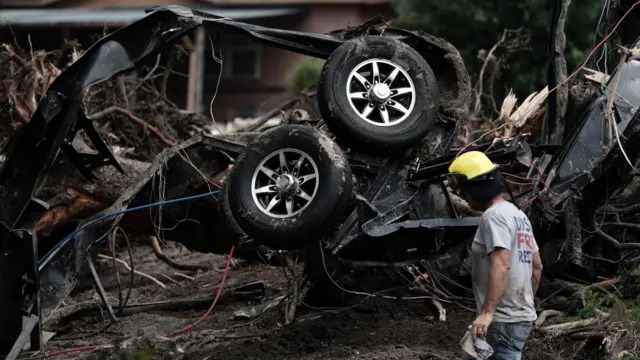 A man in a yellow helmet walks past the mangled wreckage of a car in a forested area