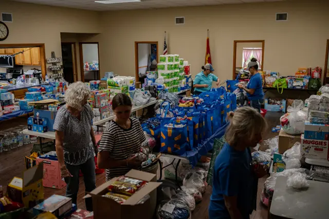 Piles of donated essentials being sorted into blue carrier bags by five women.