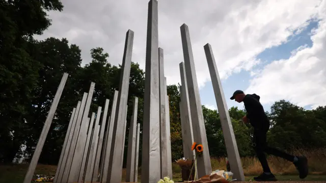 A man in a baseball cap approaches the tall thin pillars of the 7/7 Memorial at Hyde Park in London