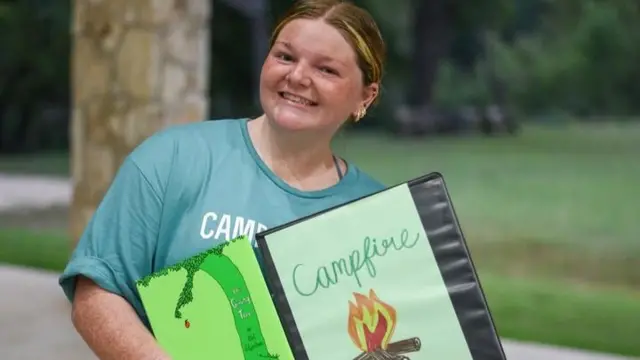 A freckled young woman smiles as she holds up a folder, the cover of which is decorated with a handdrawn campfire