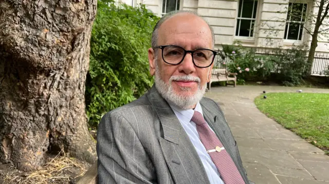 George Psaradakis in a mauve tie, grey suit and black glasses sits on a bench in a park with a tree trunk behind him.