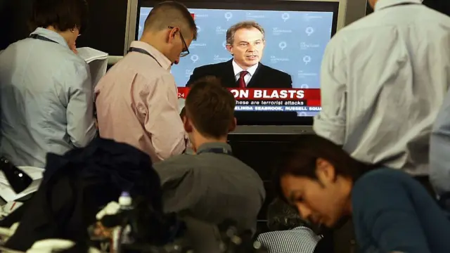 Journalists watch a live broadcast of then-Prime Minister Tony Blair addressing the nation following the 7 July 2005 London bombings. The screen headline reads “LONDON BLASTS” as Blair confirms the attacks were acts of terrorism.
