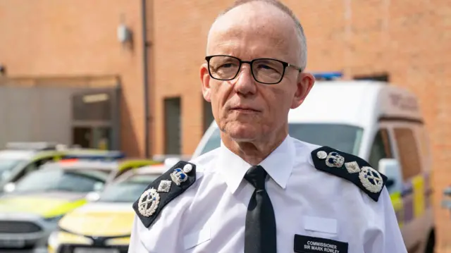 Sir Mark Rowley is seen standing in a car park with police vehicles behind him.