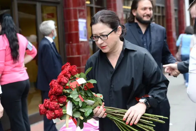 Gill Hicks is seen in black holds a bouquet of red roses outside a London Underground station, during a 2015 memorial event. Other attendees stand nearby, some wearing black ribbons in remembrance.
