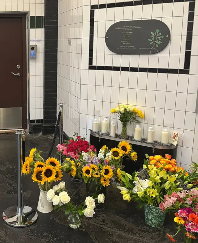 Memorial display at Edgware Road station with a plaque, candles, and floral tributes including sunflowers, roses, and lilies, marking the 7 July 2005 bombing.