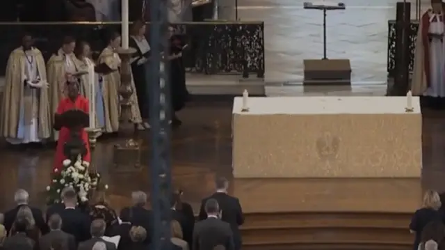 A woman wearing read stands behind a lectern, to the left of an altar.