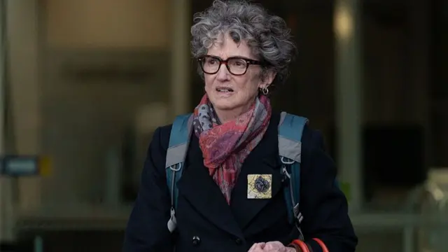 Nanette Rogers, who has grey permed hair and black spectacles, walks out of the court. She is wearing a pink scarf and navy coat, carrying a backpack.