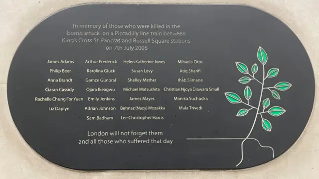 A black memorial plaque honours the victims of the 7 July 2005 bombing on a Piccadilly line train between King's Cross St. Pancras and Russell Square. It lists the names of those killed, alongside a symbolic tree design, with the words: “London will not forget them and all those who suffered that day.”