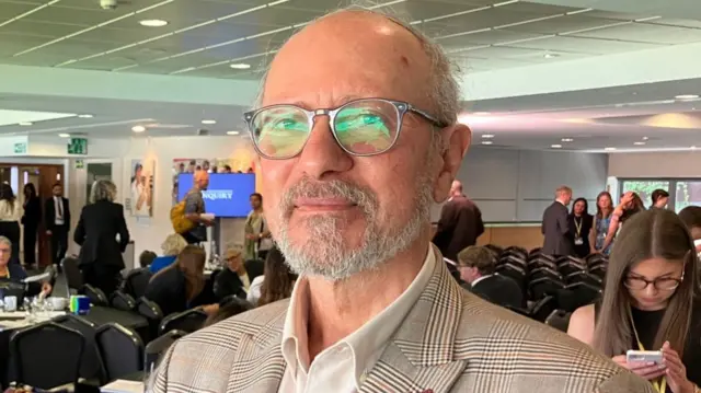 Sami Sabet looks at the camera. He is wearing a checkered blazer, white shirt, and glasses. He has a grey beard.