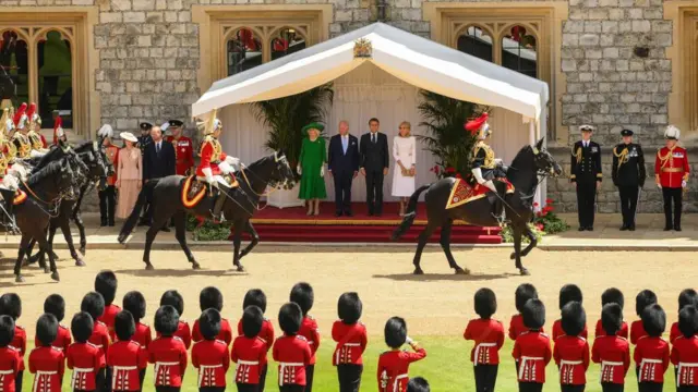 Royals stood with Macrons in a tent watching soldiers as carriages driven past them