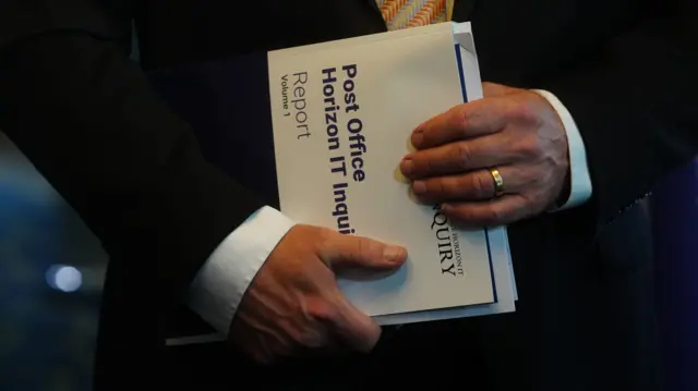 Man holding a copy of the report in his hands. he is holding it sideways in front of him. All that is visible of him is his torso.