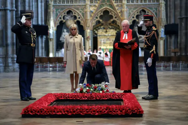 Macron lays a wreath of white, red, and blue flowers at the Tomb of the Unknown Warrior. His wife, Brigitte Macron, stands next to him. Two men are stood in military uniform on either side of the tomb doing a salute. The sub-dean of Westminster Abbey, Reverend David Stanton, is stood next to Macron.