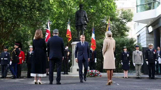 Starmer, his wife, and Brigitte Macron stand with their backs turned to the camera. Macron walks back towards them after laying a wreath at the statue