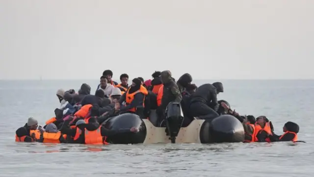 A small dinghy boat in the water with a large amount of people on it. Many people are in the water around the boat and reaching for people to pull them up