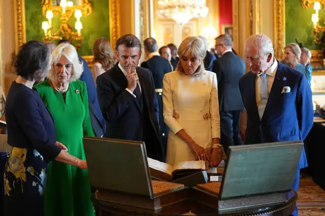 Queen Camilla, Emmanuel Macron, Brigitte Macron, and King Charles stand looking at items in boxes.