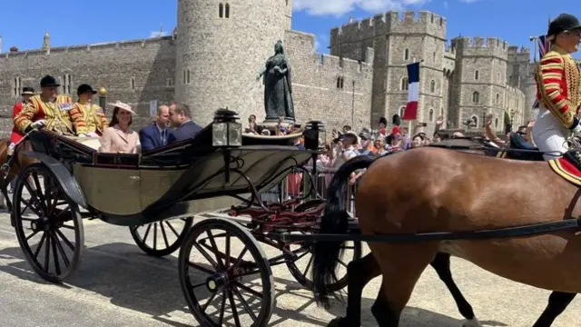 A carriage with the Prince and Princess of Wales passes by the crowd with the castle in the background