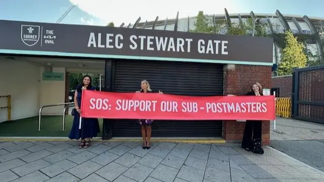 Three postmasters hold up a red sign outside the Alec Stewart Gate outside of the Oval cricket ground