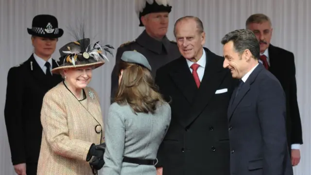 Queen Elizabeth, in a cream coloured jacket, wears a hat with feathers coming out of it and she shakes the hand of French First lady Carla Bruni-Sarkozy. To the Queen's left is her late husband, Prince Philip, who looks on smiling. French President Nicolas Sarkozy stands to the right of the group and smiles as his wife is greeted by the Queen