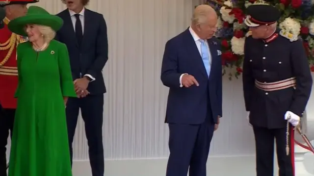 Camilla looking to one side speaking while Charles stands near her talking to a soldier