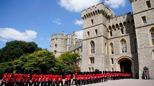 Windsor Castle with soldiers marching into it through an arch. They are wearing red uniforms and black fur caps.