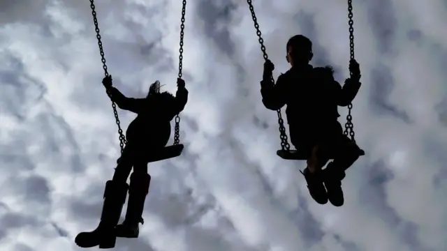 Two children are seen as silhouettes playing on swings in a park