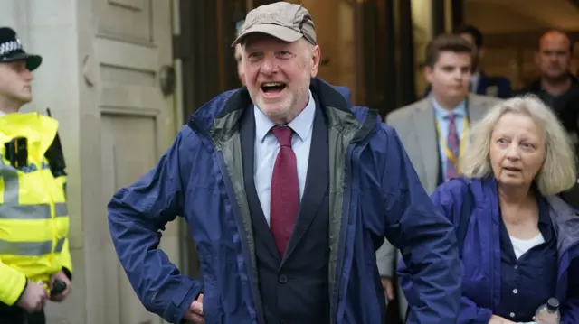 Post Office campaigner Alan Bates wears a suit, blue anorak and green cap. He's leaving a building and a police officer stands to his right-hand side.