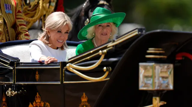 Brigitte Macron smiles while sat in a carriage next to Queen Camilla. Brigitte is wearing a white dress and Queen Camilla is wearing a green dress and hat. Both women have blonde hair.