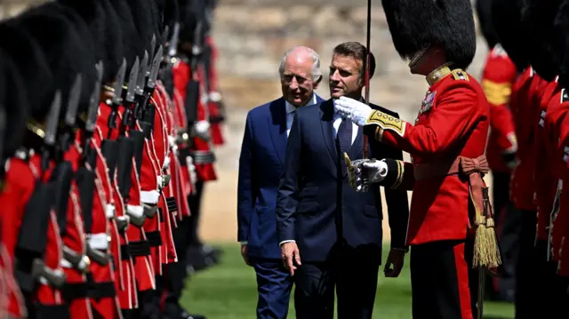 Macron and King Charles walk along in front of a row of soldiers wearing red uniforms and black fur caps. The soldiers are holding guns. Macron and King Charles are both wearing navy suits. King Charles has white hair and Macron has brown hair.