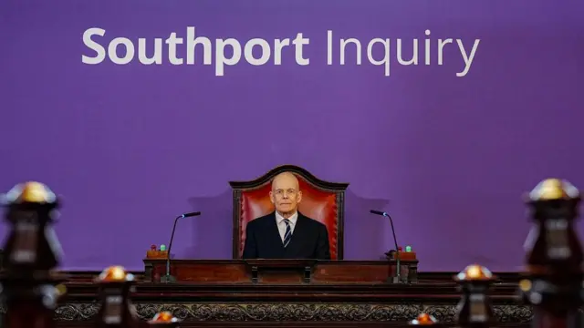 A man in a suit sits at a very imposing desk in the back of a fancy room. Behind him the wall is purple and says Southport Inquiry in big letters