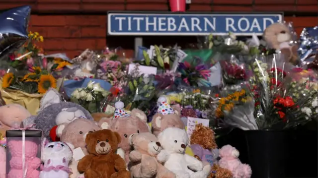 Flowers and teddy bears lay on the road in front of a sign reading Tithebarn Road in Southport