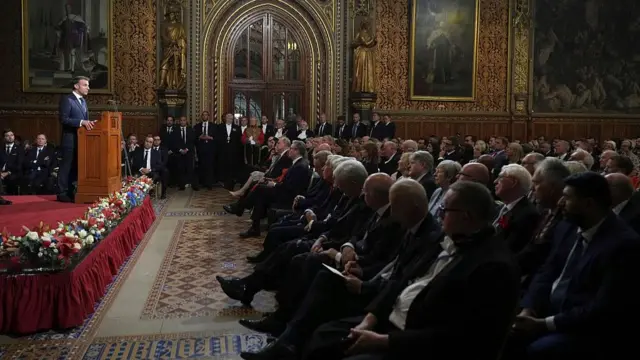 Emmanuel Macron addresses MPs inside of Westminster. He stands at a podium, whilst the crowd sits and listens