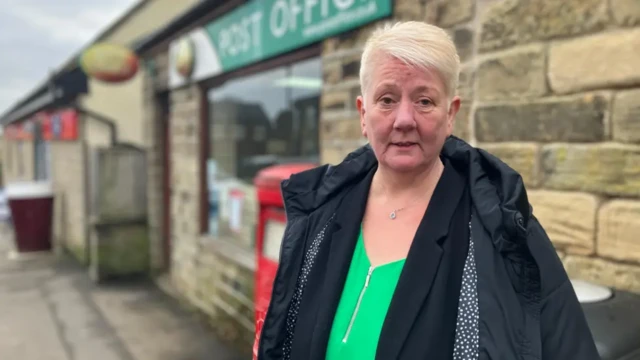 Alison Hall pictured outside a Post Office wearing a green shirt and black jacket. She has blond short hair.