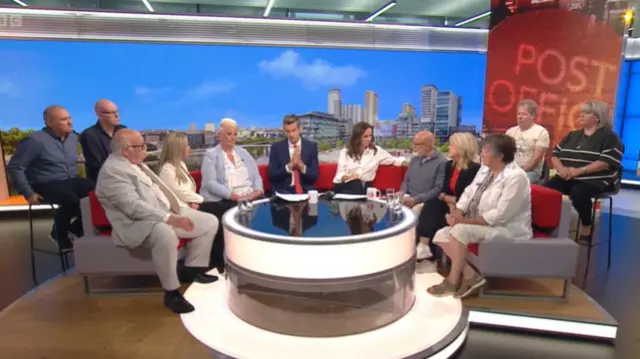 Wide shot of BBC Breakfast studio showing presenters and guests sitting down on a grey and red couch, a screen showing the Salford skyline in the background