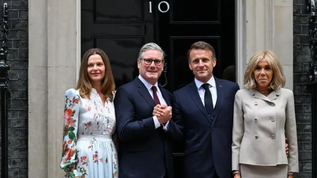 Left to right: Victoria Starmer stands next to Keir Starmer, who holds hands with Emmanuel Macron, who stands next to Brigitte Macron