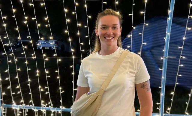 Maisie smiling into the camera. She’s wearing a white t-shirt and a beige over the shoulder bag. She has blonde hair with darker roots. The background is a load of fairy lights.