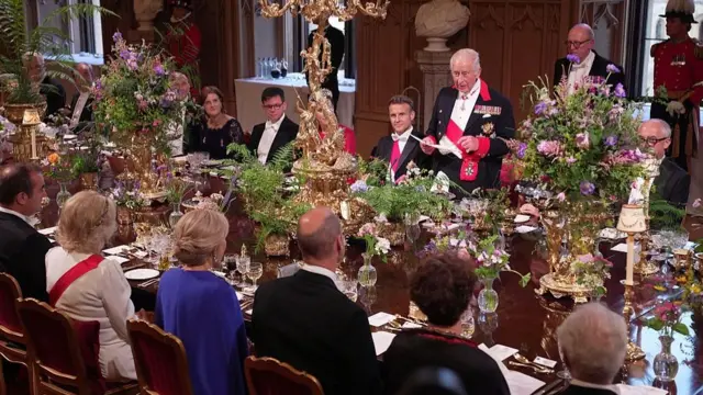 Emmanuel Macron sits next to Princess of Wales to his right and on his right is King Charles III, who addresses a large state banquet. The King is standing, whilst everyone else is seated at the table.