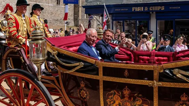 Emmanuel Macron rides in a horse-drawn carriage alongside King Charles III. The French president waves to onlookers, who snap pictures on their phones.
