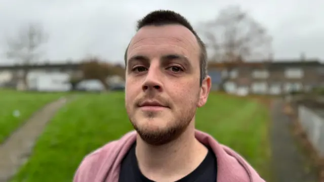 A man in a pink hoodie stands in front of a row of houses behind a green on an overcast day.