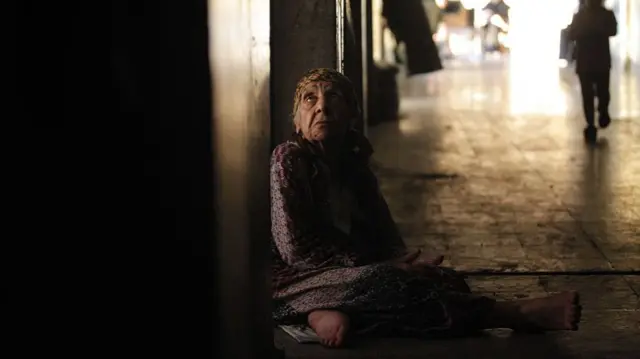 An elderly Palestinian woman sits against a wall in a corridor a a building where she is sheltering