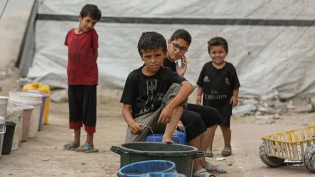 Four boys under tarpaulin shelters sit on water buckets