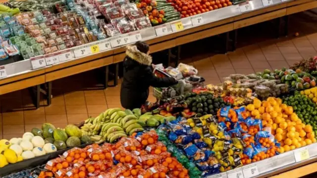 Woman in coat with fur hood pushing a full trolley through an aisle of groceries in supermarket.