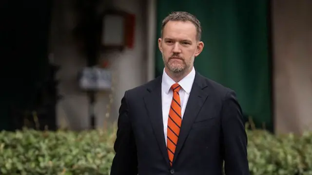 Greer walking in front of a hedge in a suit and tie, looking serious