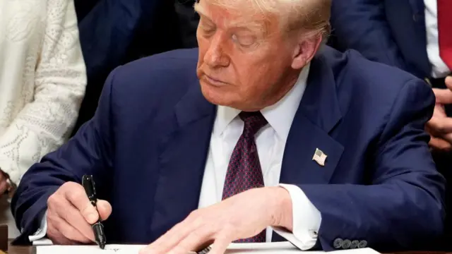 A close up of Donald Trump signing paperwork at the Oval Office desk